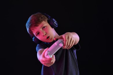 Cute little boy in headphones playing video game with controller on black background