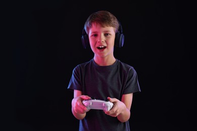 Cute little boy in headphones playing video game with controller on black background