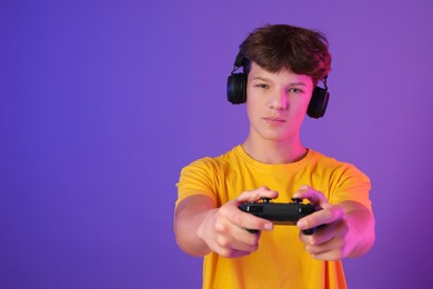 Teenage boy in headphones playing video game with controller on purple background. Space for text