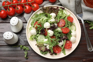 Photo of Tasty salad with arugula, lettuce, mozzarella cheese, vegetables and fork on wooden table, flat lay