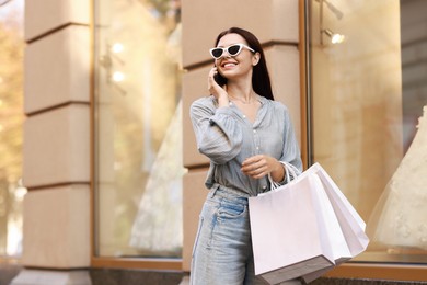 Happy woman with shopping bags talking on smartphone outdoors, space for text