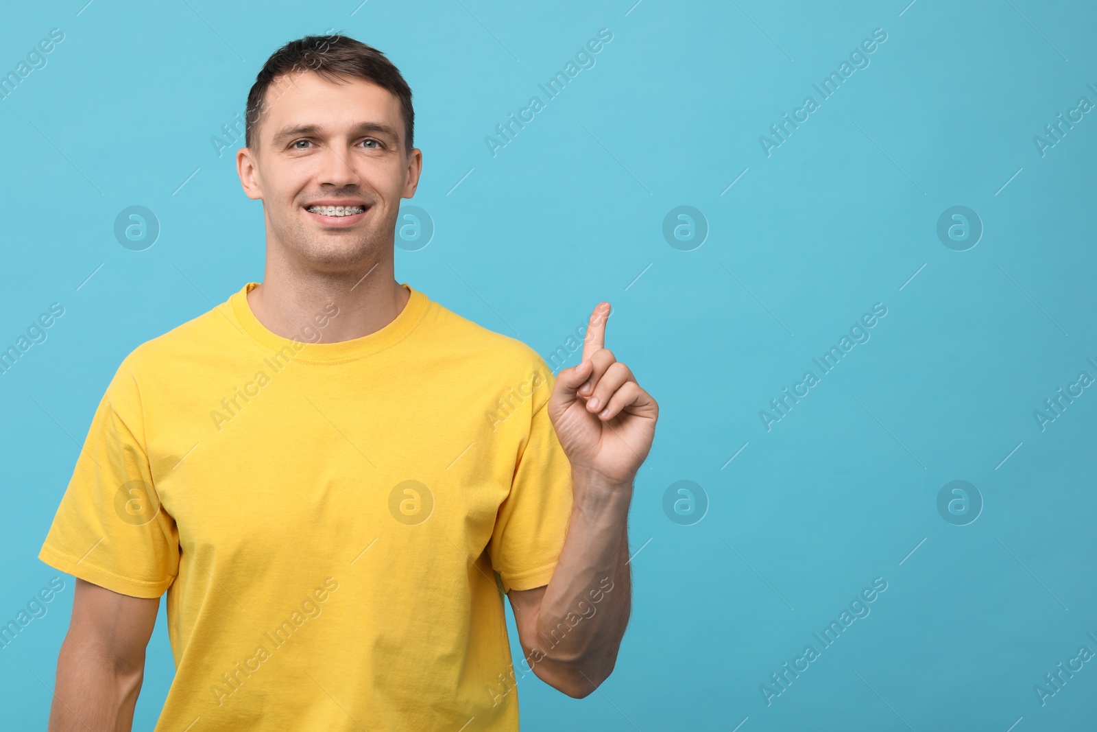 Photo of Smiling man with dental braces pointing at something on light blue background. Space for text