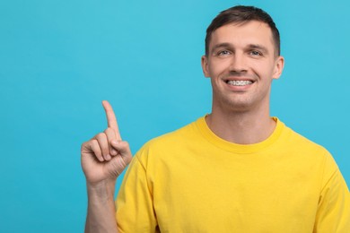 Photo of Smiling man with dental braces pointing at something on light blue background. Space for text