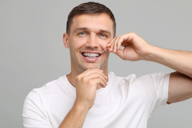 Smiling man with braces cleaning teeth using dental floss on grey background