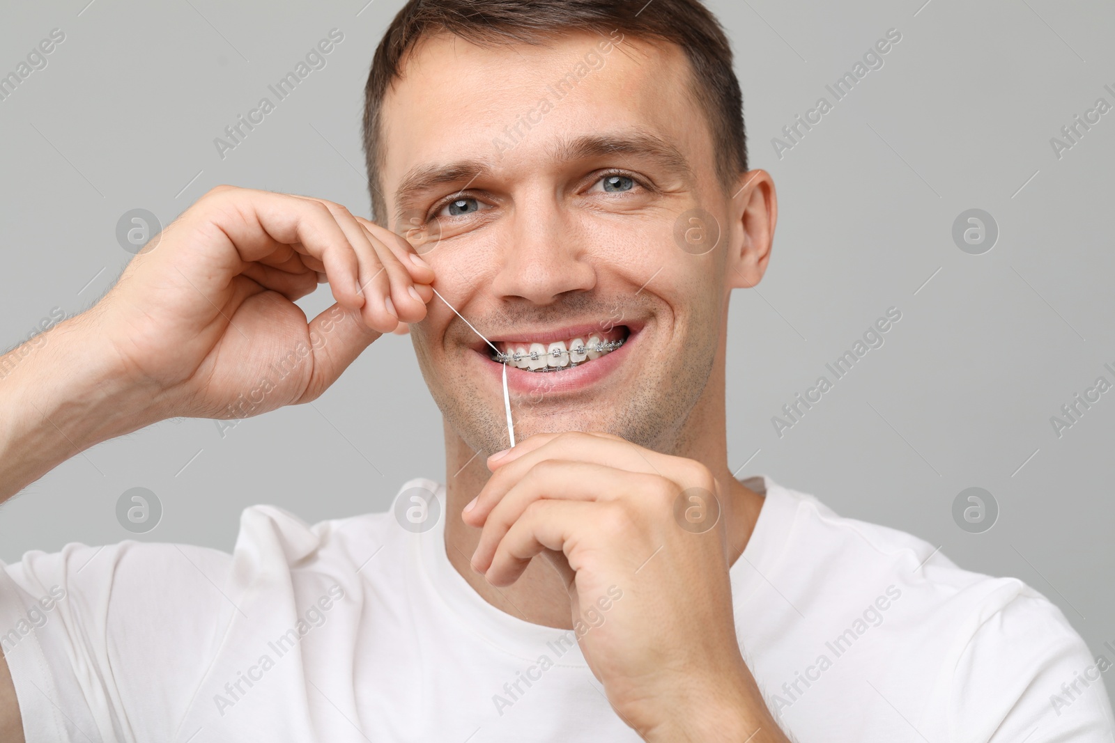 Photo of Smiling man with braces cleaning teeth using dental floss on grey background