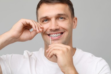 Smiling man with braces cleaning teeth using dental floss on grey background