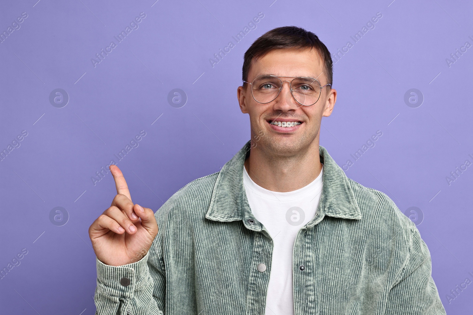 Photo of Smiling man with dental braces pointing at something on violet background. Space for text