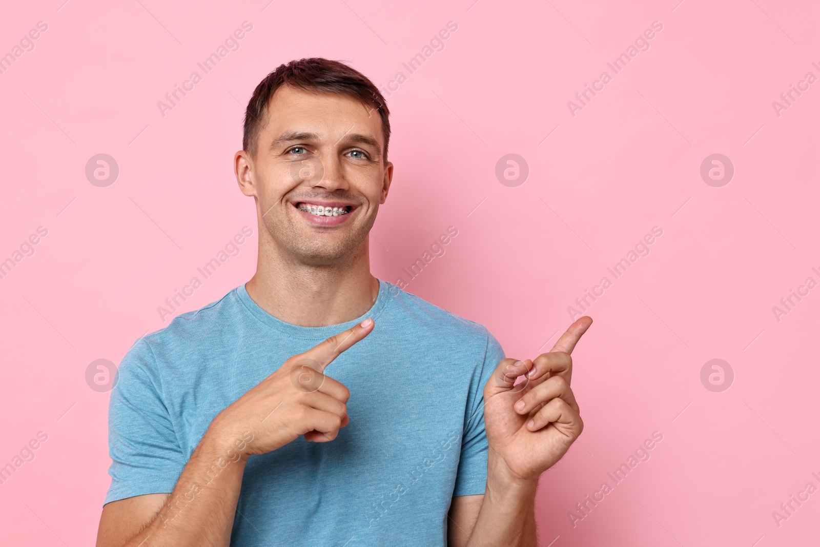 Photo of Smiling man with dental braces pointing at something on pink background. Space for text