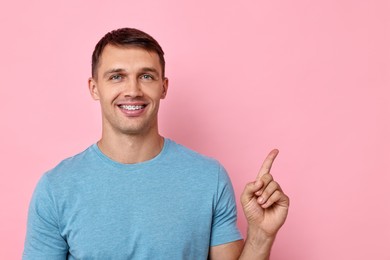 Photo of Smiling man with dental braces pointing at something on pink background. Space for text