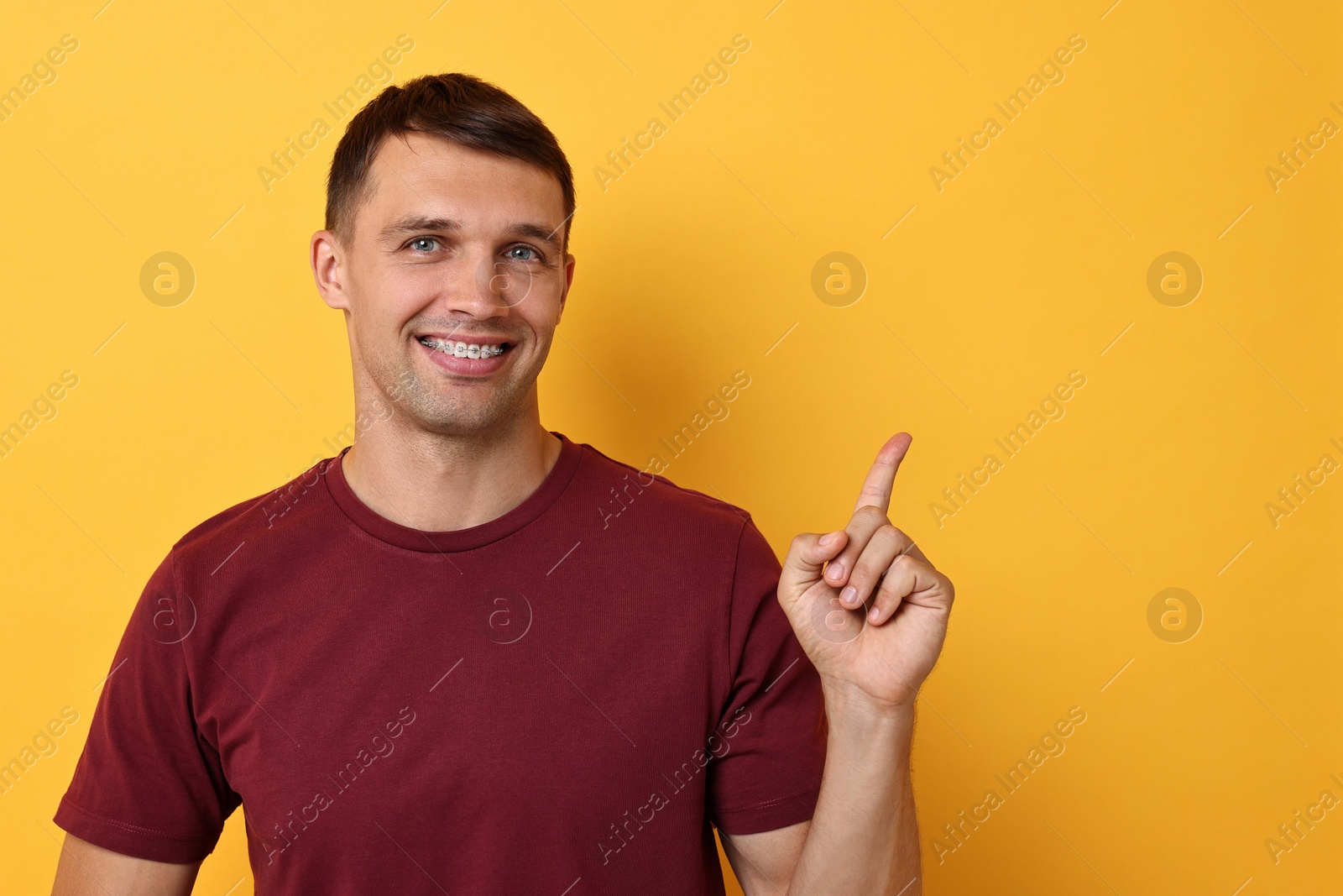 Photo of Smiling man with dental braces pointing at something on yellow background. Space for text