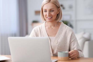 Happy middle aged woman with cup of drink using laptop at table indoors