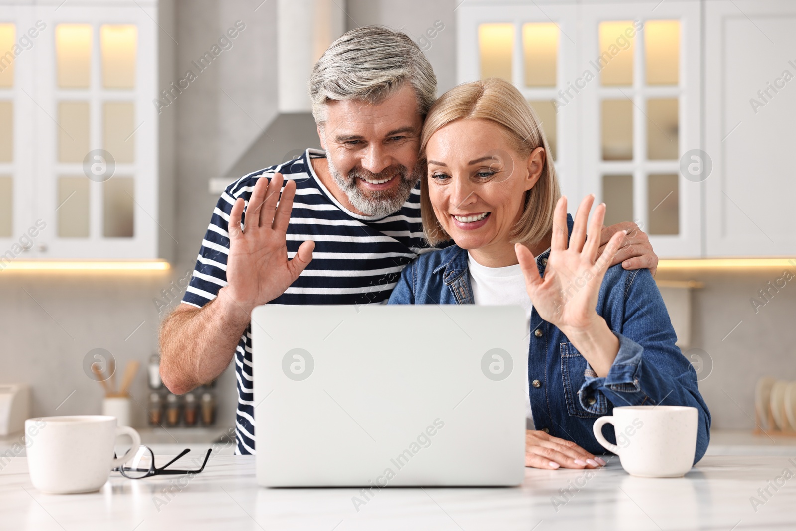 Photo of Happy middle aged couple having video chat via laptop at white marble table in kitchen