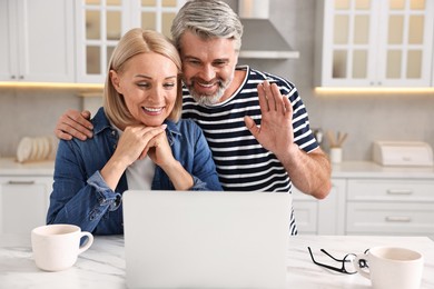 Happy middle aged couple having video chat via laptop at white marble table in kitchen