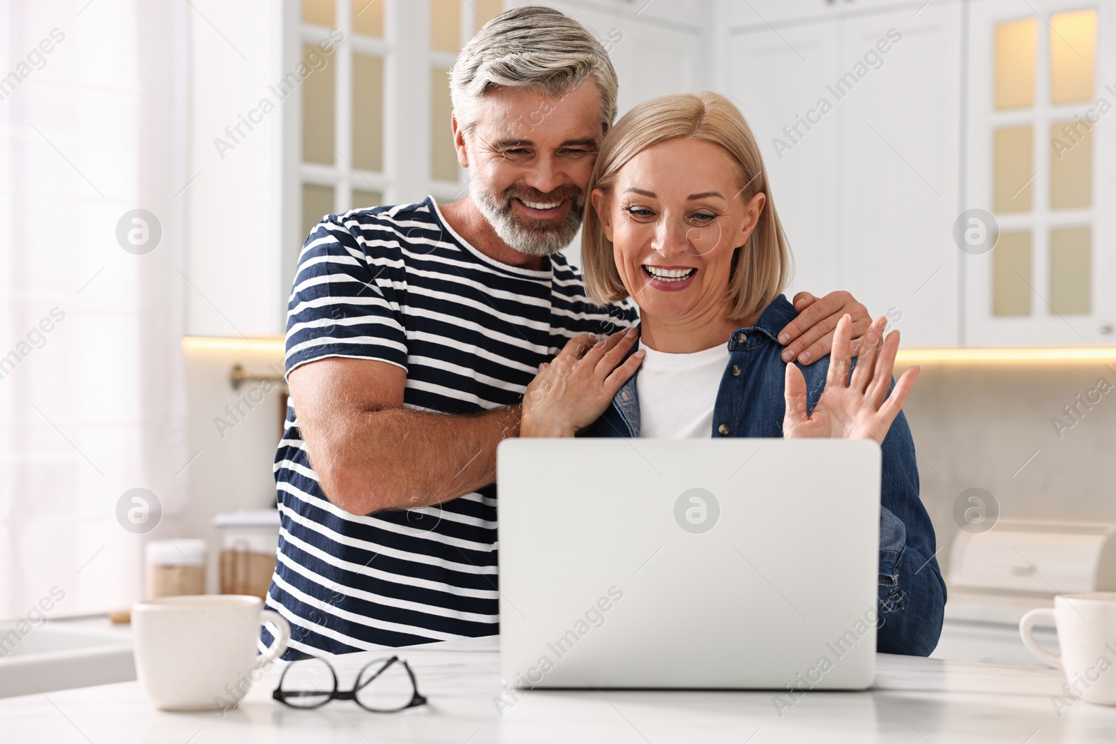 Photo of Happy middle aged couple having video chat via laptop at white marble table in kitchen