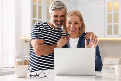 Happy middle aged couple having video chat via laptop at white marble table in kitchen