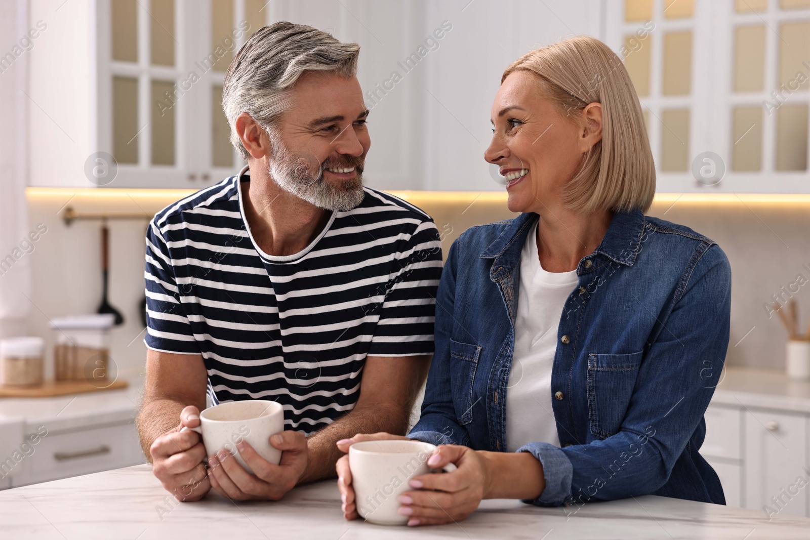 Photo of Happy middle aged couple with cups of drink at white marble table in kitchen