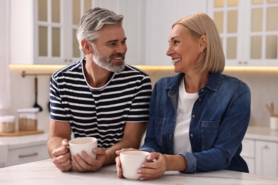 Happy middle aged couple with cups of drink at white marble table in kitchen