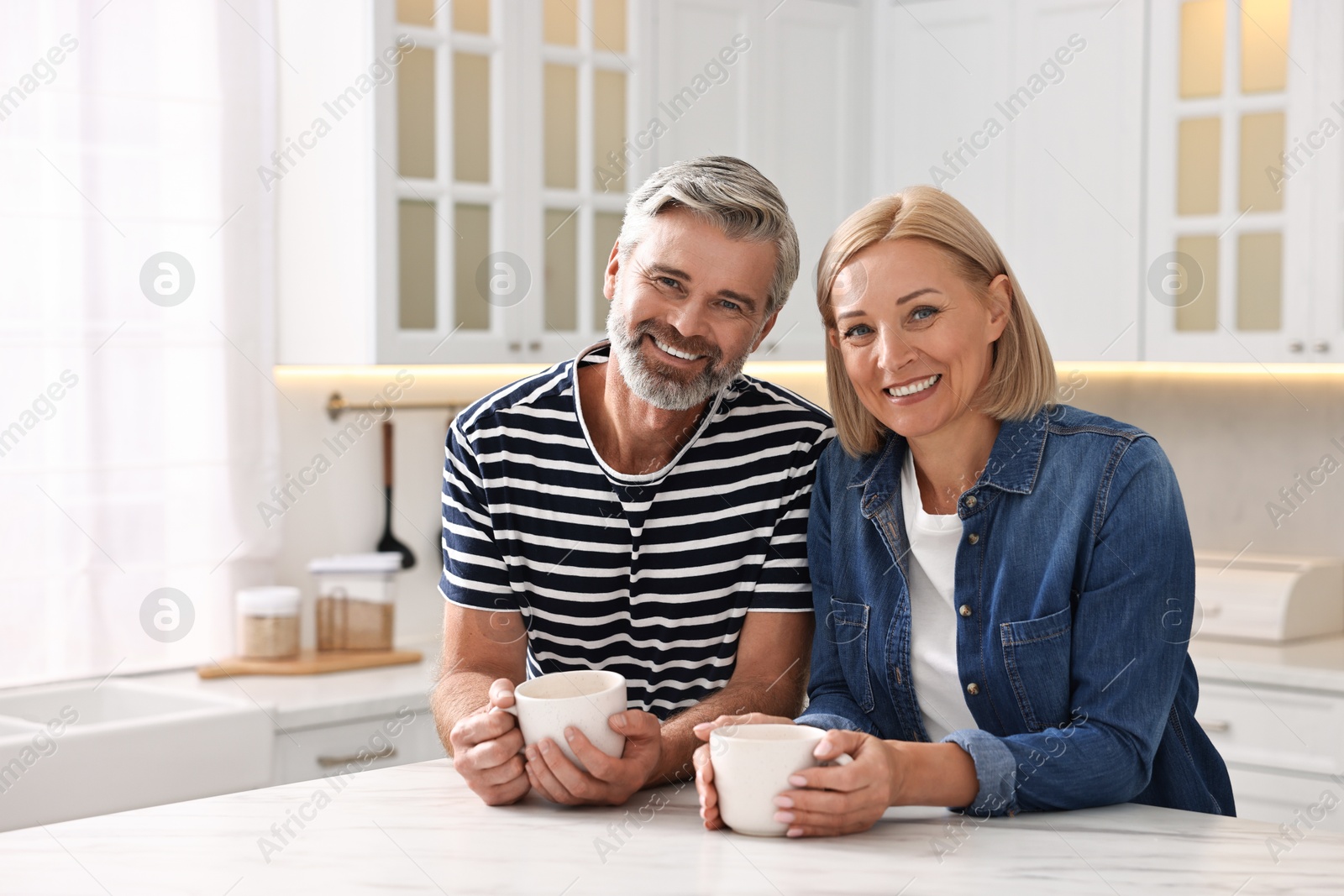 Photo of Happy middle aged couple with cups of drink at white marble table in kitchen