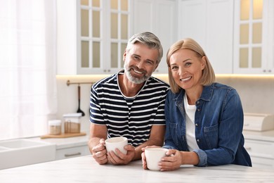 Happy middle aged couple with cups of drink at white marble table in kitchen