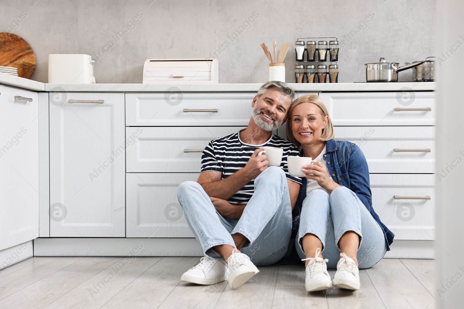 Photo of Happy middle aged couple with cups of drink on floor in kitchen, space for text