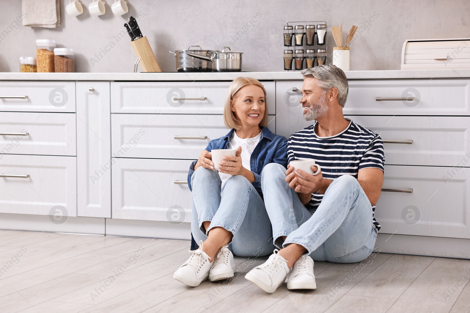 Photo of Happy middle aged couple with cups of drink on floor in kitchen, space for text