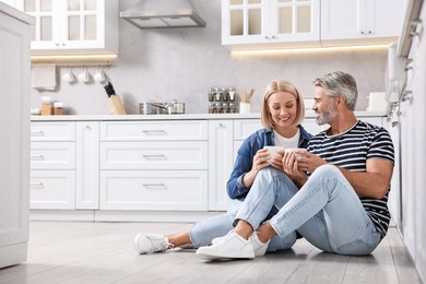 Happy middle aged couple with cups of drink on floor in kitchen, space for text