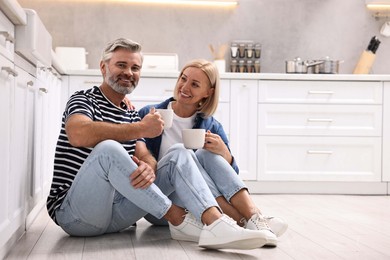 Photo of Happy middle aged couple with cups of drink on floor in kitchen, space for text