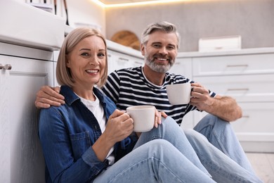 Happy middle aged couple with cups of drink on floor in kitchen