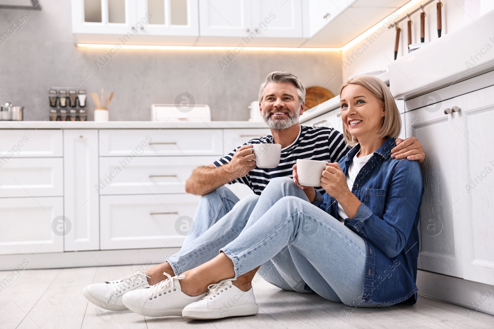 Photo of Happy middle aged couple with cups of drink on floor in kitchen, space for text