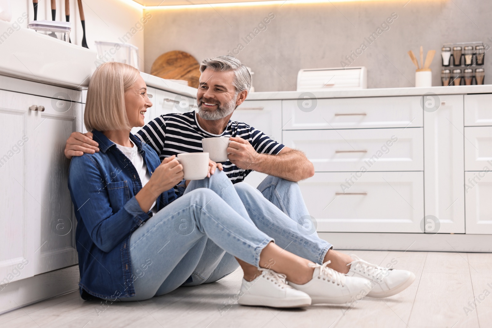 Photo of Happy middle aged couple with cups of drink on floor in kitchen