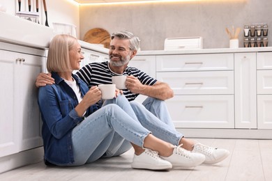 Happy middle aged couple with cups of drink on floor in kitchen