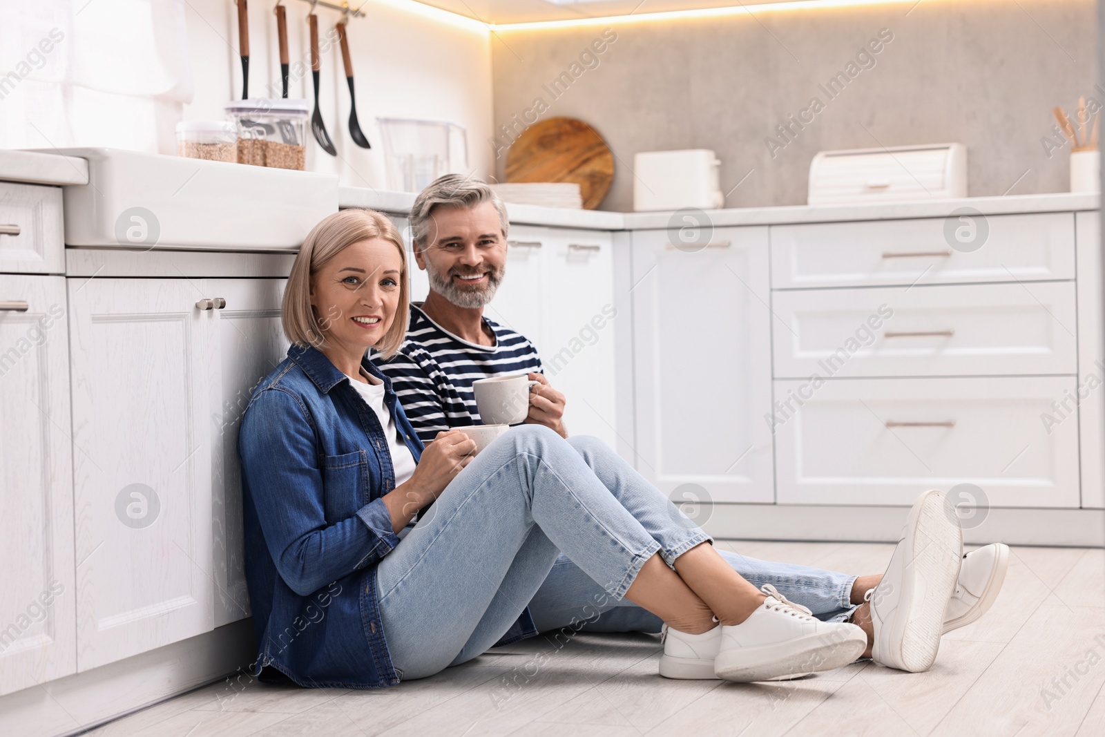 Photo of Happy middle aged couple with cups of drink on floor in kitchen