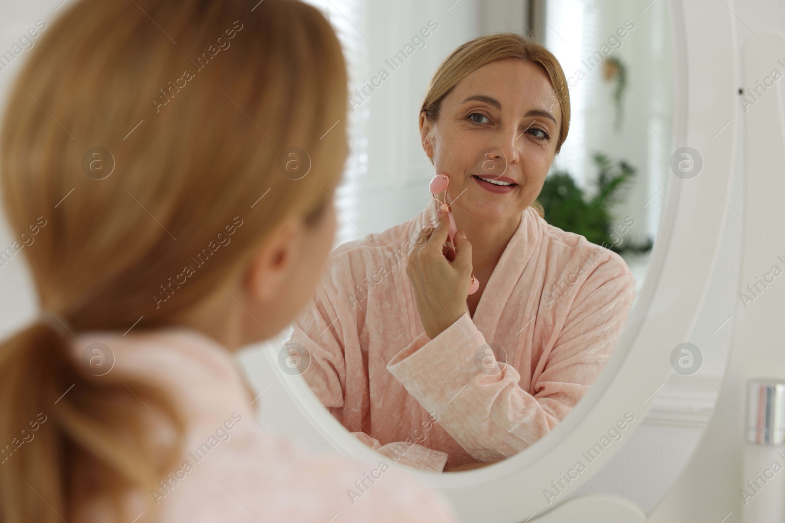 Photo of Smiling woman doing facial self massage with roller near mirror in bathroom