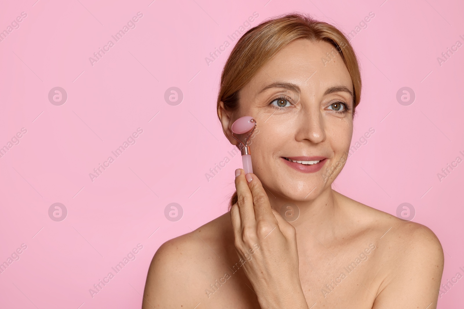 Photo of Smiling woman doing facial self massage with roller on pink background. Space for text