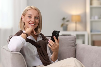 Smiling middle aged woman with smartphone on armchair at home