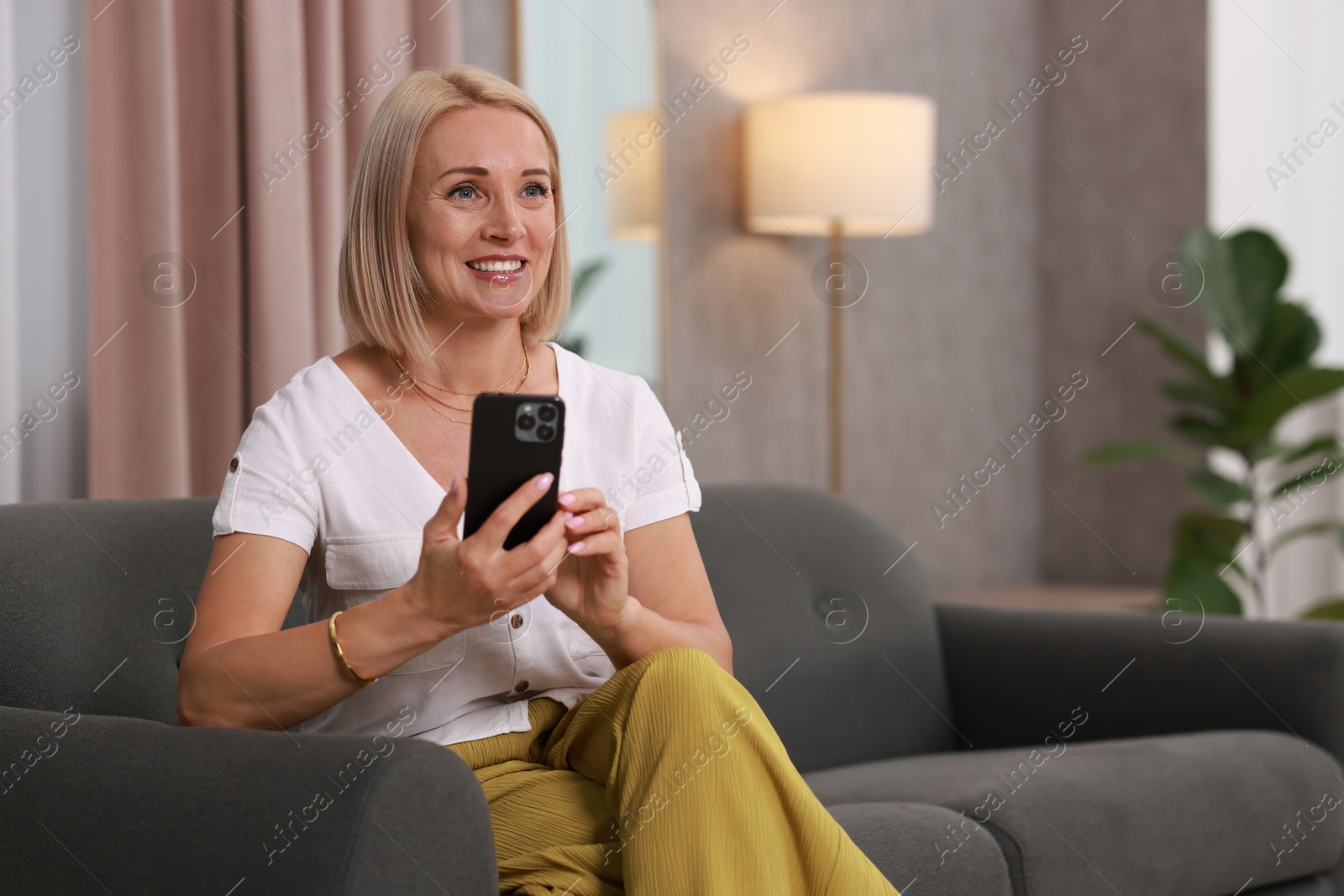 Photo of Portrait of smiling middle aged woman with smartphone on sofa at home. Space for text