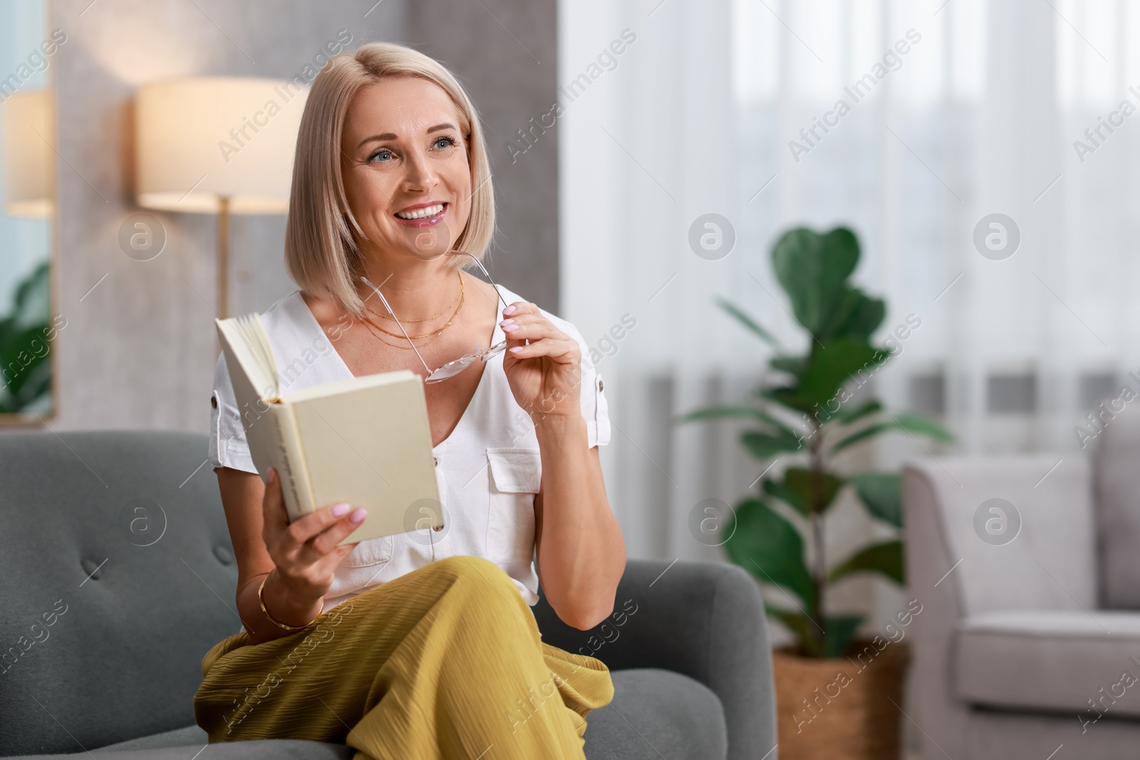 Photo of Portrait of smiling middle aged woman with glasses reading book on sofa at home. Space for text