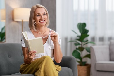 Portrait of smiling middle aged woman with glasses reading book on sofa at home. Space for text