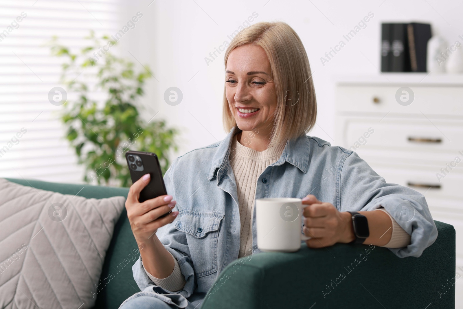 Photo of Smiling middle aged woman with cup of hot drink using smartphone at home