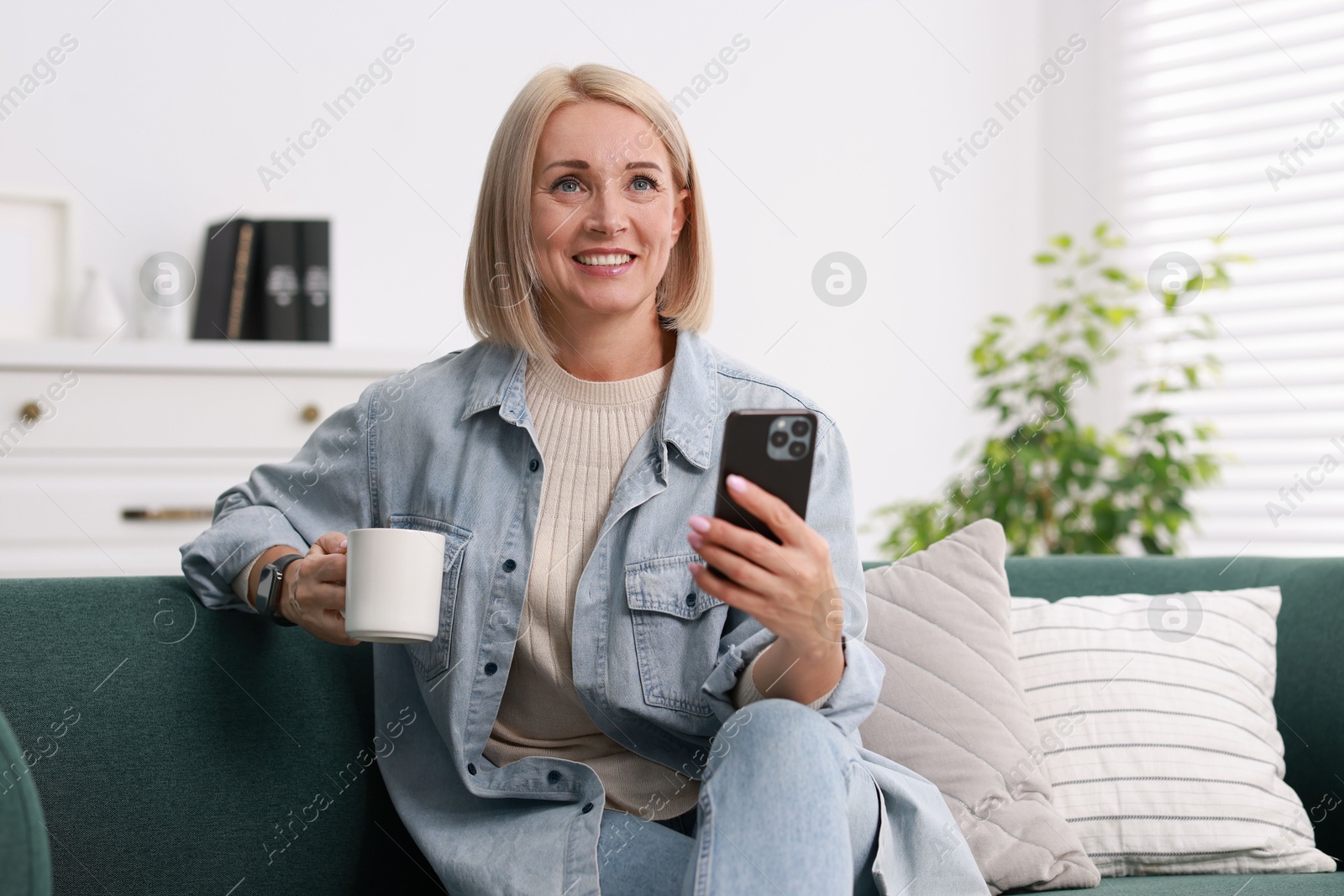 Photo of Smiling middle aged woman with cup of hot drink using smartphone at home