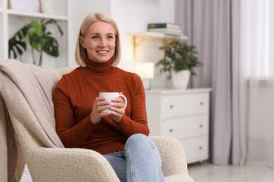 Smiling middle aged woman with cup of hot drink on armchair at home. Space for text