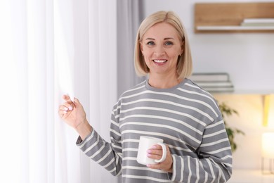 Smiling middle aged woman with cup of hot drink near window at home