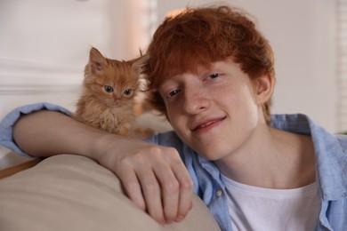 Photo of Redhead teenage boy with cute ginger kitten on sofa indoors