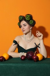 Photo of Young woman with peppers in her hair, fruits and vegetables at green table on orange background