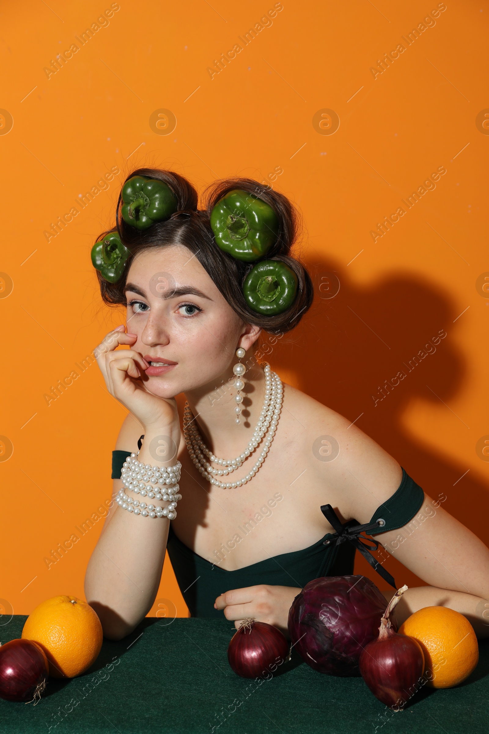 Photo of Young woman with peppers in her hair, fruits and vegetables at green table on orange background