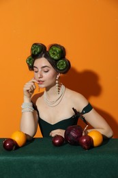 Young woman with peppers in her hair, fruits and vegetables at green table on orange background