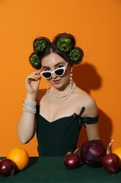 Young woman with peppers in her hair, sunglasses, fruits and vegetables at green table on orange background