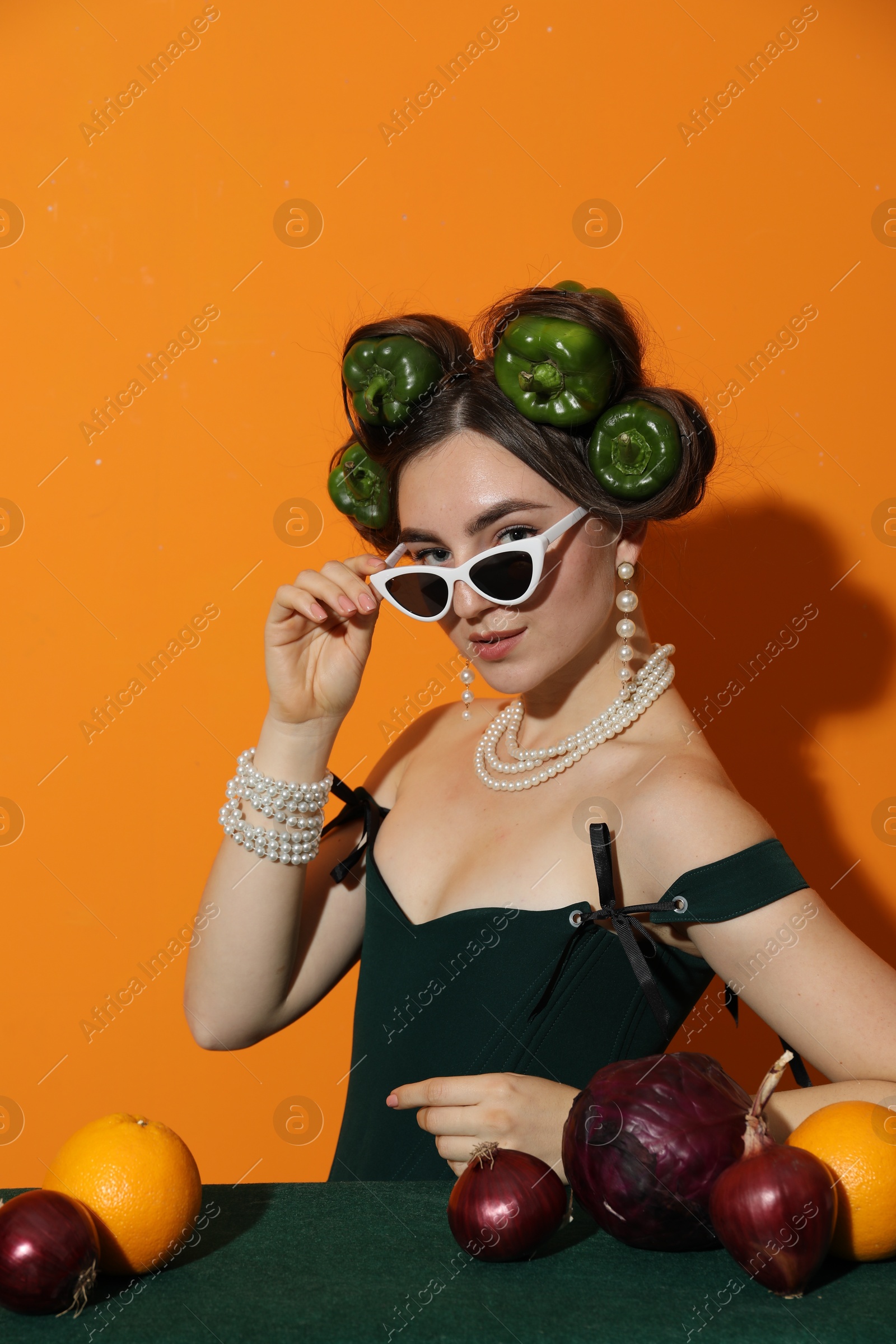 Photo of Young woman with peppers in her hair, sunglasses, fruits and vegetables at green table on orange background