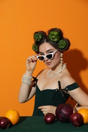 Young woman with peppers in her hair, sunglasses, fruits and vegetables at green table on orange background