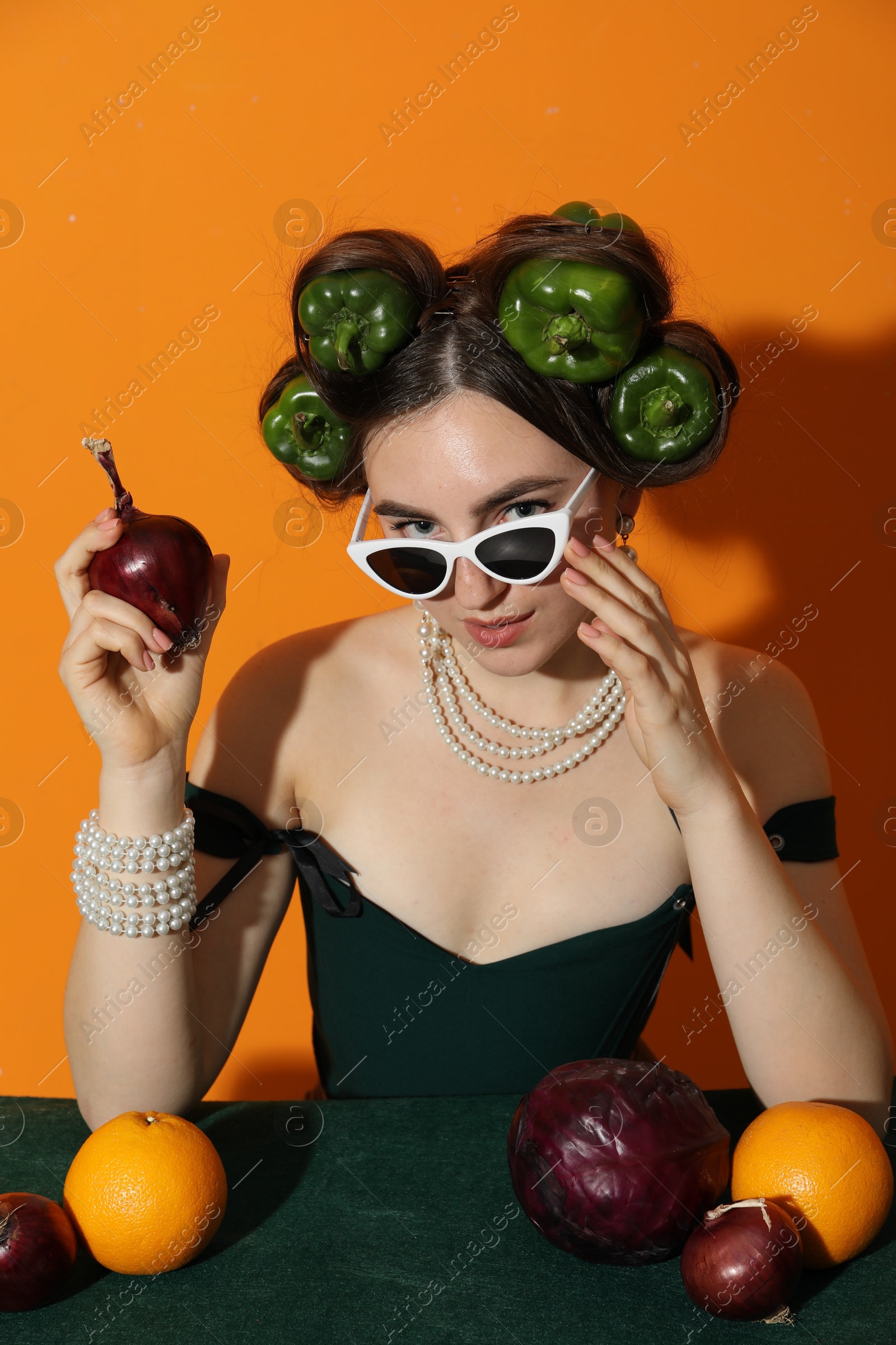 Photo of Young woman with peppers in her hair, sunglasses, fruits and vegetables at green table on orange background